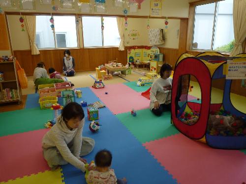 写真：3組の親子がおもちゃで遊んでいる様子。おひさまルームの床にはプレイングマットが敷かれ、ブロックやおままごとセット、ハウス型ボールプールなどのおもちゃがあります。
