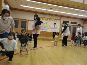 写真：しまとりゲームに参加し音楽に合わせて職員と一緒に手をつないでラインの上を歩いているひよこ組の子どもたち