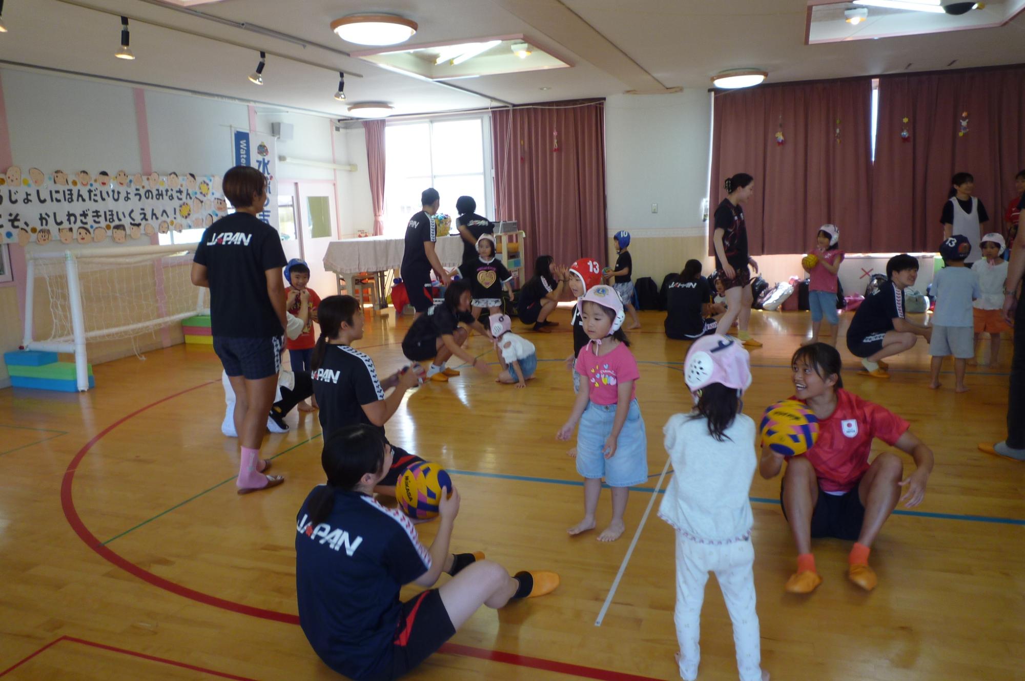 写真：水球競技用の帽子を被った5歳女児が、水球選手からボールの扱い方を教えてもらっているところ。
