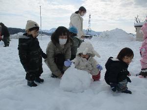 写真：スノーウェアを着た0歳児の男の子2人と女の子1人が、園庭に積もった雪に触れて遊んでいるところ