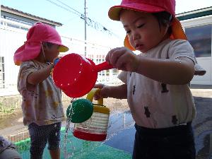 写真：手桶に汲んだ水をじょうろに移したち、じょうろから流れる水で遊ぶ1歳児2人