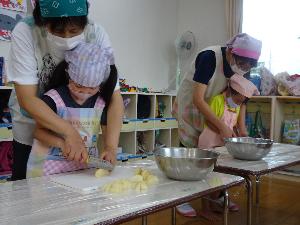 写真：先生に後ろから手を添えてもらいながら、包丁でジャガイモを切る2人の5歳児