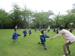 写真：両手を広げた保育士に向かって、子どもたちが広い芝生でかけっこをしている様子