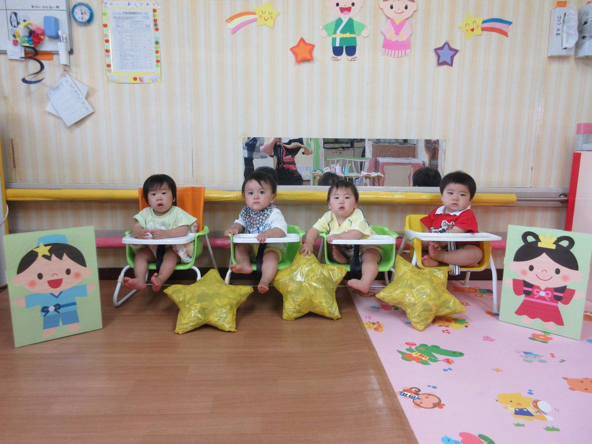 写真：七夕のゲームで使った星型の風船やや織姫、彦星の看板と並んで椅子に座る0歳児4人