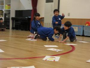 写真：自分で作ったA4サイズほどのカルタで遊んでいるところ。遊戯室に取り札が並べられ、読み上げ度同時に勢いよく取りに行きます。