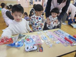 写真：郵便ごっこの抽選会で、机の上に並んだ景品の玩具を選んでいる3人の園児