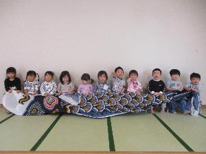 2歳児11人が横一列に座り、大きな黒いこいのぼりを広げて持っている写真