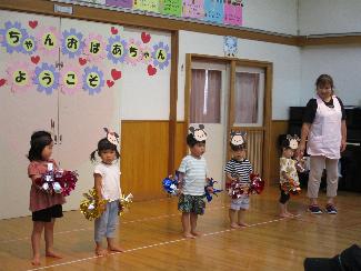 写真：祖父母お招き会で踊りを披露する2歳児。頭には動物のお面をつけ、手にポンポンをもって一列に並んでいます