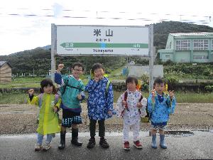 写真：米山駅の看板の前で、雨具を着た5人の子どもたちが並んでいます。どの子も笑顔で、電車に乗るのを楽しみにしている様子です。