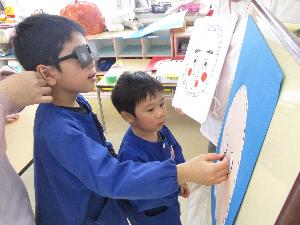 写真：目隠しをした5歳児が、ペアになった3歳児のアドバイスで福笑いをしています。