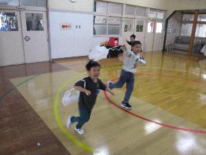 写真：ビニール袋に紐を括り付けた凧をあげて、楽しむ子どもたち。お遊戯室を笑顔で走り回っています。