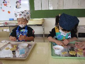 写真：エプロンと三角巾をつけた2人の子どもが、トレーに乗っているクッキー生地にトッピングしている様子