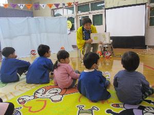 写真：5人の子ども達が横一列に並び、黄色い法被を着たスタッフに絵本を読んでもらっている様子