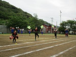 写真：小学校の校庭で1列に並び、黒い耳付き帽子をかぶり、旗やポンポンを持って踊る4人の園児の様子
