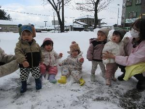 写真：ひよこ組の園児が園庭で初めての雪遊びを経験しているところ。雪を触ったり、雪の上に立ったりしています