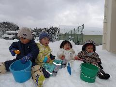 写真：ひよこ組の子ども達が雪の上に座り、スコップやバケツを使って雪遊びをしているところ