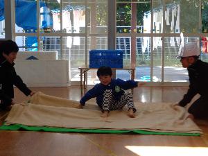 写真：毛布の端を持った2人の職員が、園児が載った毛布を揺らし、地震体験をしています。