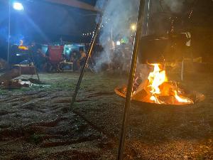 写真：夜のキャンプエリア。手前に燃えている薪が写り、後ろにキャンプチェアに腰掛け団らんしてる人たちが見えます。