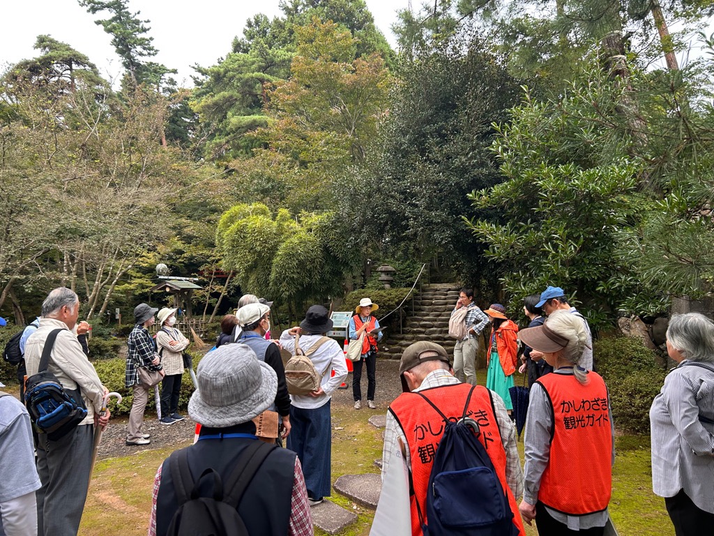 写真：オレンジ色のベストを着たボランティアガイドが、松雲山荘内で解説しています。松が多く植えられ、奥には木村茶道美術館へ向かう階段があります。