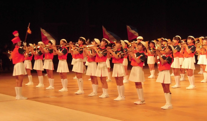 写真：白いベレー帽と赤い上着、白いスカートや半ズボンの揃いの衣装で、アルフォーレの舞台で演奏する小学校のマーチングバンド