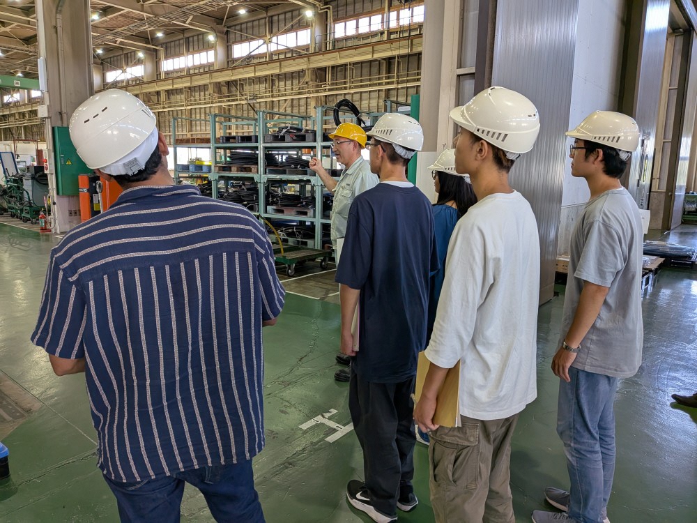 写真：工場内を見学する学生たち。工場の奥にはたくさんの太いケーブルなどが並んでいます