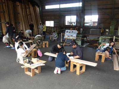 写真：木材加工場で、参加した親子が、材木にしるしをつけたり、のこぎりを使って切ったりする作業をしています