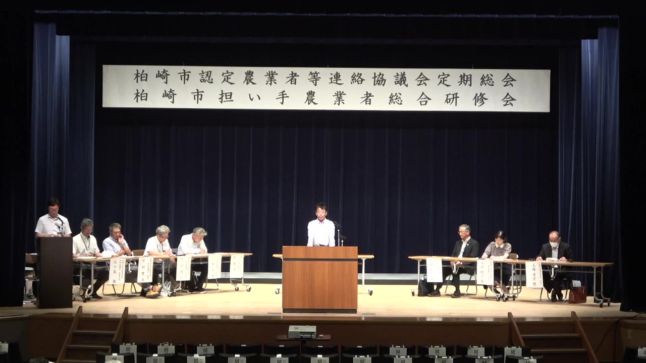 写真：総会の様子。ステージに役員らが並び、提案された議事を協議しています。