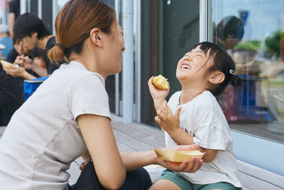 写真：お母さんと向かい合って座る女の子が、片手におにぎりを持ち、幸せそうな笑顔をみせています。