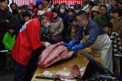 写真：大きなマグロを解体する男性2人。その周りにはたくさんのお客さんが集まり見学しています