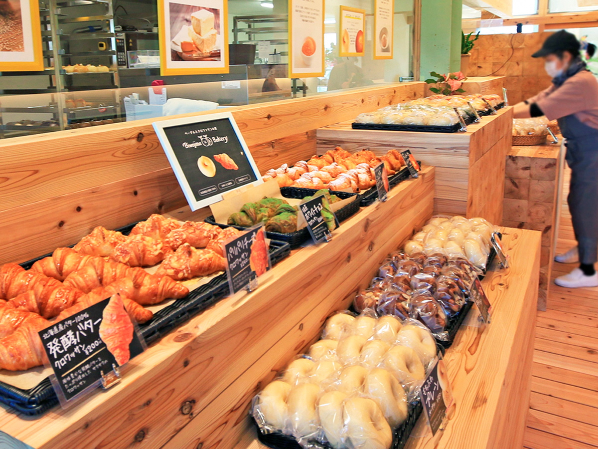 写真：木目調の店内にベーグルやクロワッサンなどいろんな種類のパンが陳列されています。店内奥はガラス張りになっており、厨房の様子が見えます。