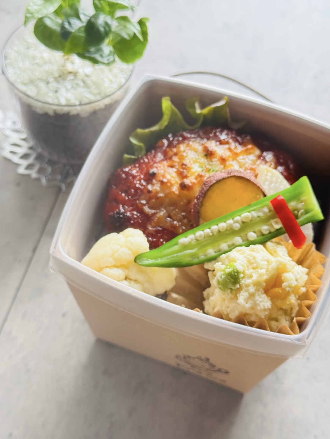 写真：かぼちゃのチーズハンバーグにカリフラワーやオクラ、サラダが添えられています。です。お弁当箱で、下段にご飯が入っている2段式のお弁当です