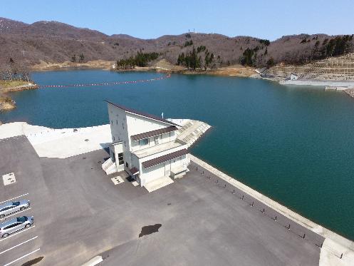 写真：写真中央に真っ白な管理棟があり、その奥に水をたたえたダム湖が写っています。