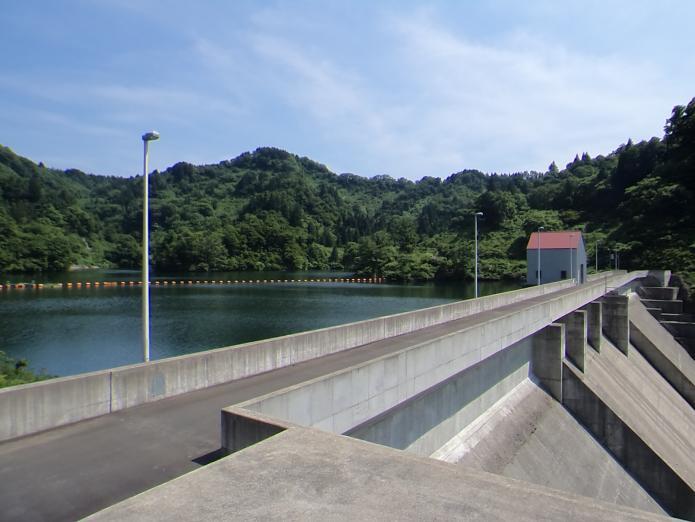 青い空と緑がきれいな山とダムの水をバックに堤体を左岸より撮影した写真