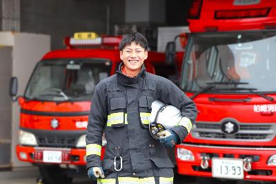 写真：ヘルメットを脇に抱え、防火衣を着用した山田さん。後ろに2台の消防車両が見えます
