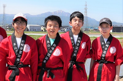 田植えに参加した東中学校2年生の生徒4人が、赤いはっぴを着て並んでいます。