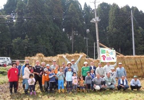 田んぼに立てられた「はさ」の前で、参加者が集合している写真