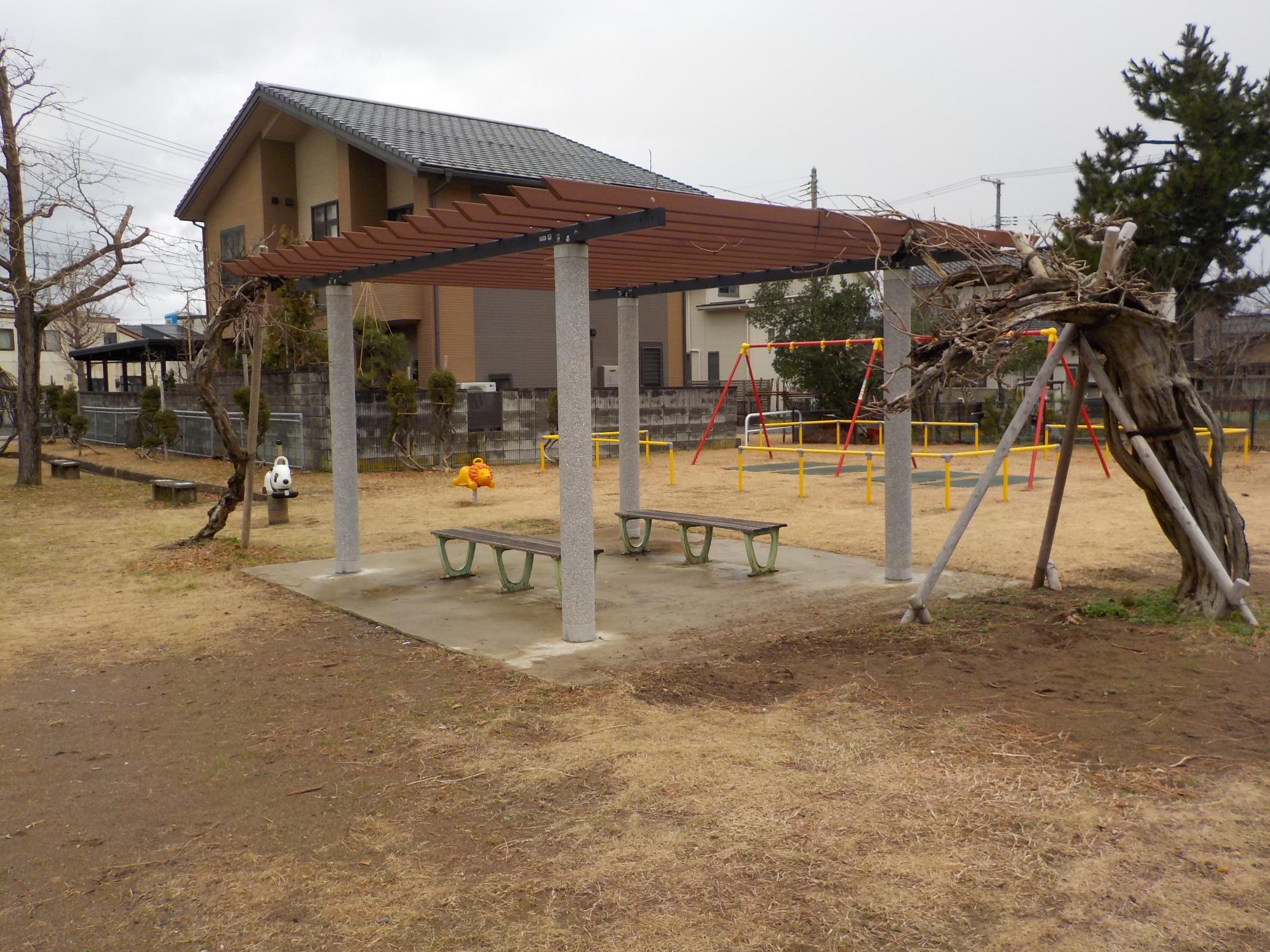 写真：パーゴラの下にベンチが向かい合わせに設置されています。その奥にスプリング型遊具とブランコがあります。