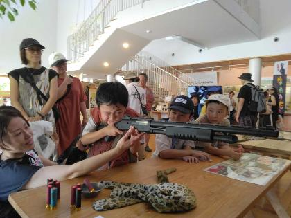 写真：射的用のライフルを持ち、片目で狙いを定めている男の子。お友達やお母さんが周りで見守っています。