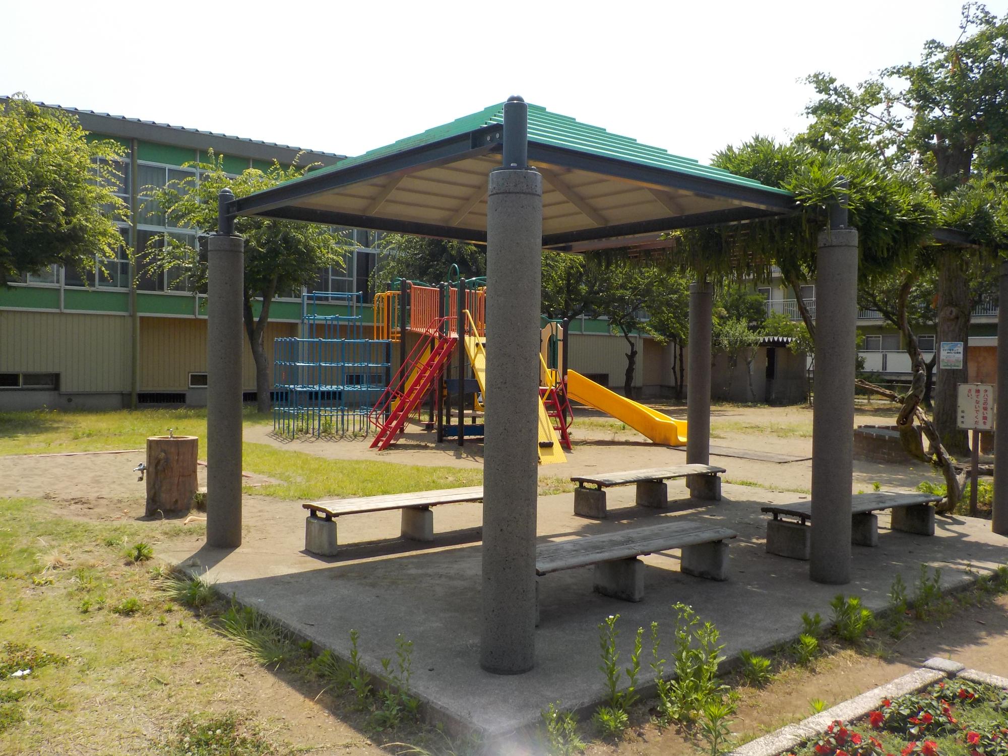 写真：手前にパーゴラとずまや、奥に滑り台のついた複合遊具があります。その奥には比角コミュニティセンターの体育館が見えます。