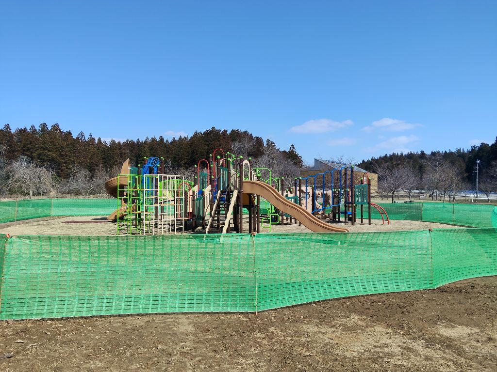 写真：広場中央に大型の複合遊具（複数の滑り台やジャングルジムなど）があります。芝生の養生中のため、遊具の周りに柵がしてあります