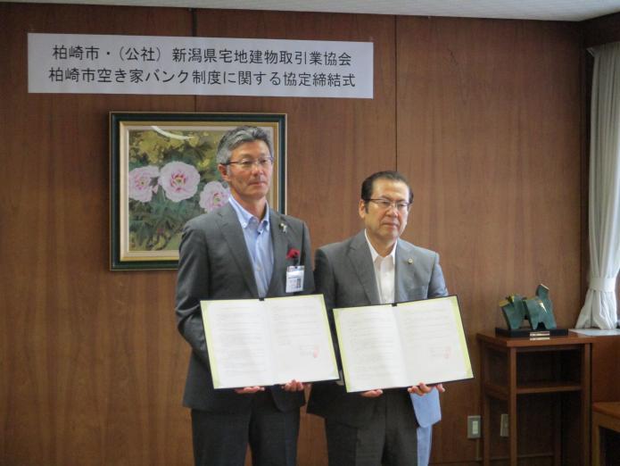 部屋の壁に飾られた花の絵画の前に立って手に協定に関する文書をもっている柏崎市長と新潟県宅地建物取引業協会の代表者の写真
