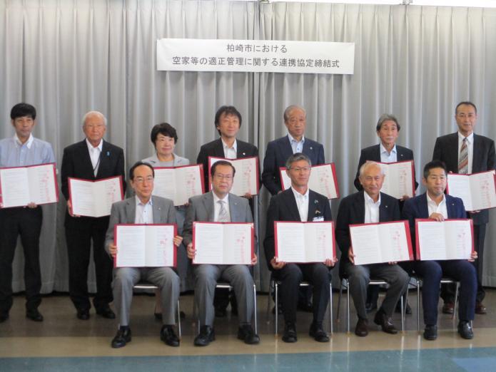 スーツ姿で椅子に腰かけている5人の男性とその後ろに立っている6人の男性と1人の女性が全員手に赤い表紙がついた文書を持っている写真
