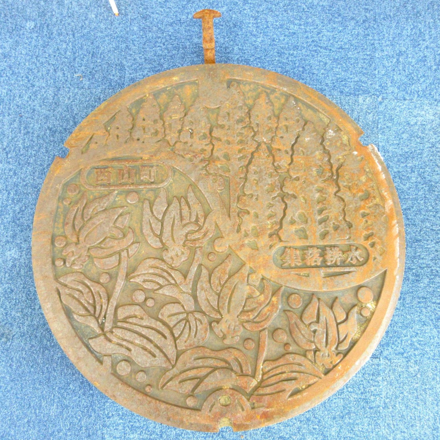 旧西山町デザインマンホール蓋の写真。上部から右側にかけて杉の木が、下部から左側にかけてカタクリの花がデザインされています。