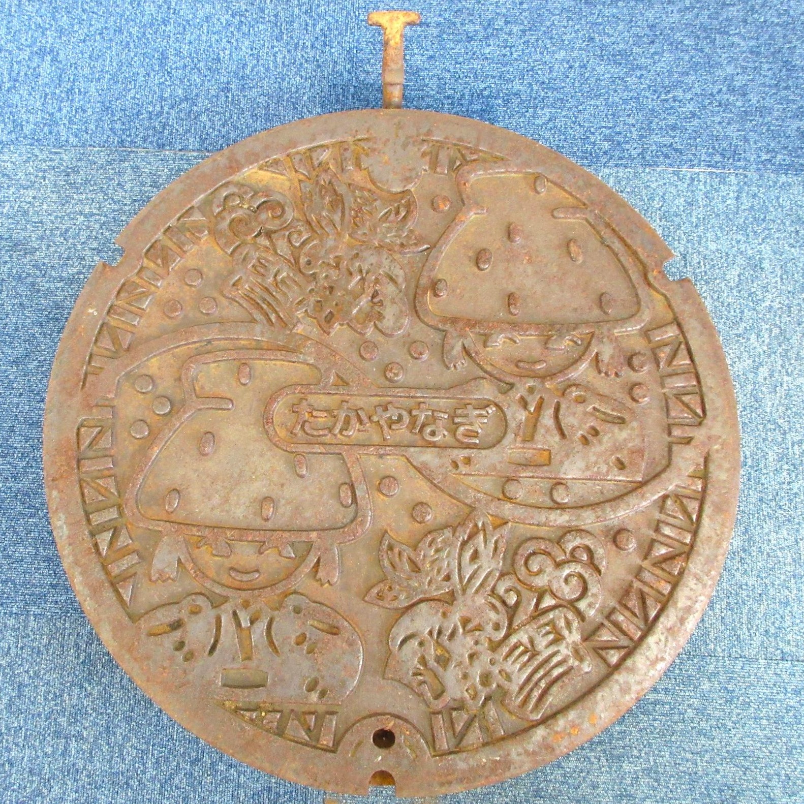 旧高柳町デザインマンホール蓋の写真。中央に高柳の文字があり、かやぶき屋根を模した髪型の女の子と山菜がデザインされています