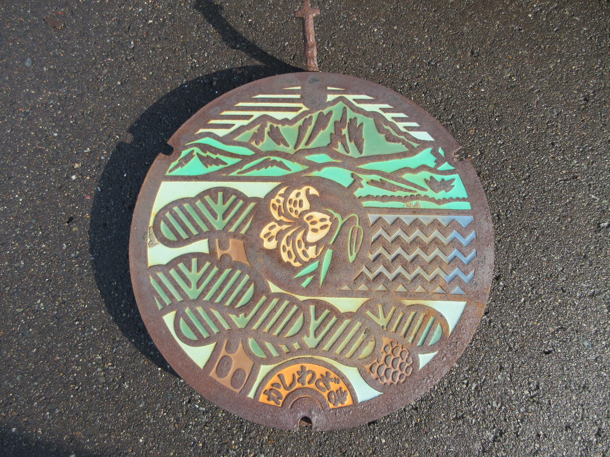 使用済みマンホール蓋の写真。中央にヤマユリ、上部から時計回りに米山、日本海の波、松が描かれています。