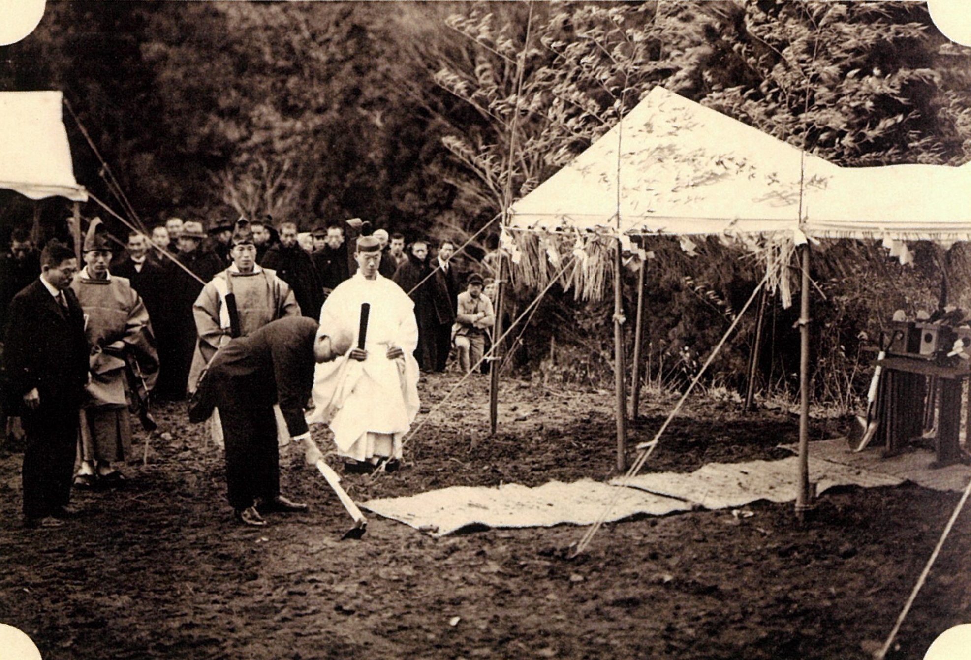 写真：川地ダムでの地鎮祭の様子。テントの4隅に竹が括り付けられ、テントの下には祭壇とお供え物があります。テントの前には2人の神主がいて、その前で男性が地面にクワを入れています。トの4隅に
