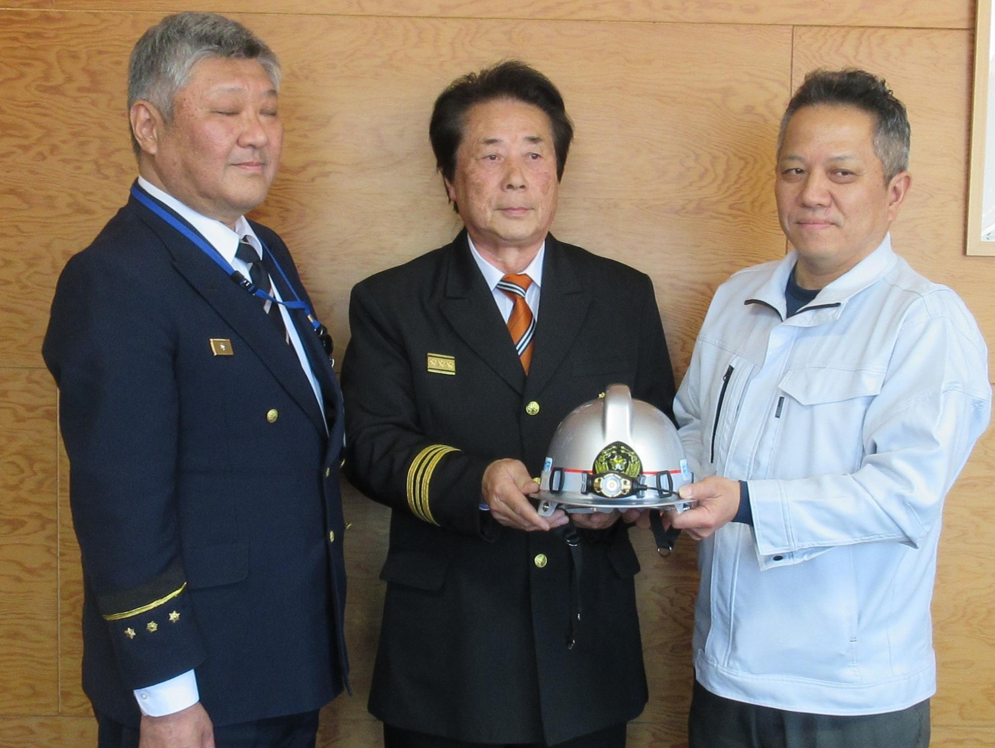 写真：左から順に浅野消防長、田村柏崎市消防団長、丸高建設高橋代表取締役が並んでいます。消防団長と高橋取締役がヘッドライトを装着したヘルメットを二人で持っています