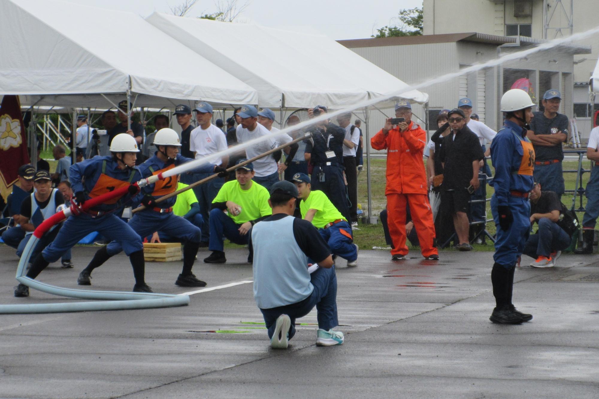 写真：指揮者が前方で火点を見据え、指揮者の後方で1番員が火点に向けて放水を行っています。また、1番員の左隣で2番員が火点に向けてホースを構えています