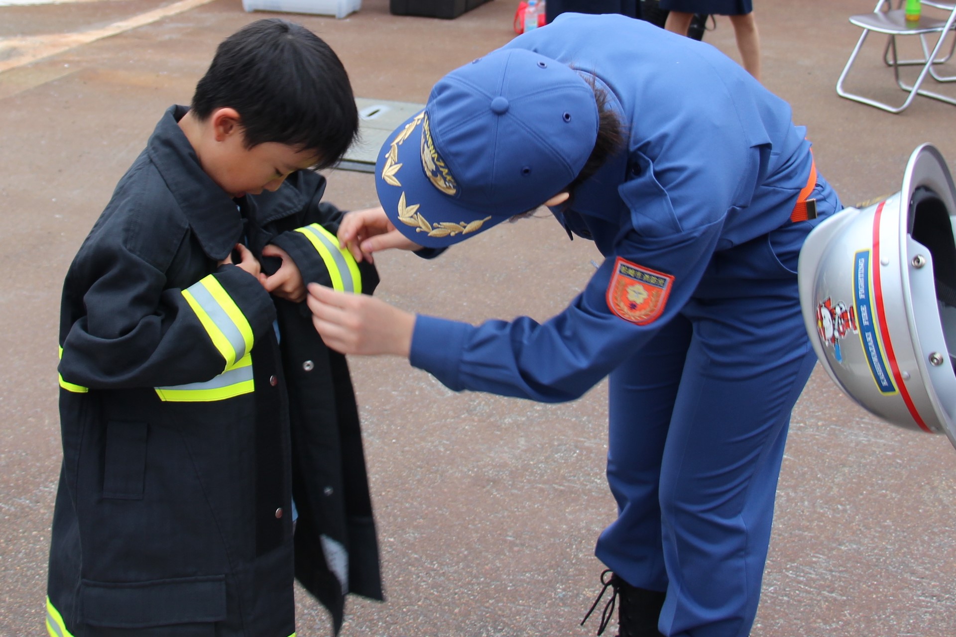 写真：子供用の防火衣を着用する男児。学生消防隊員がボタンを留めるのを手伝っています