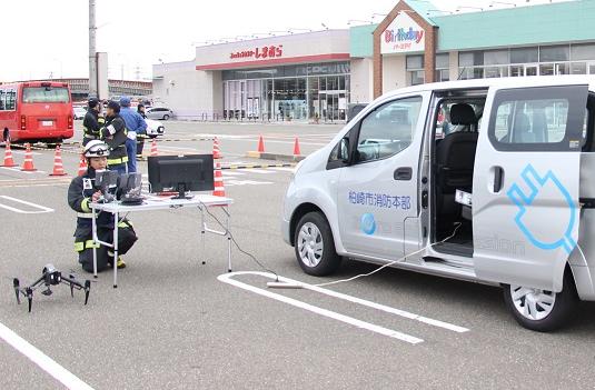 スーパーの駐車場でドローンの映像を映し出すモニター機材を電気自動車につなげているところの写真